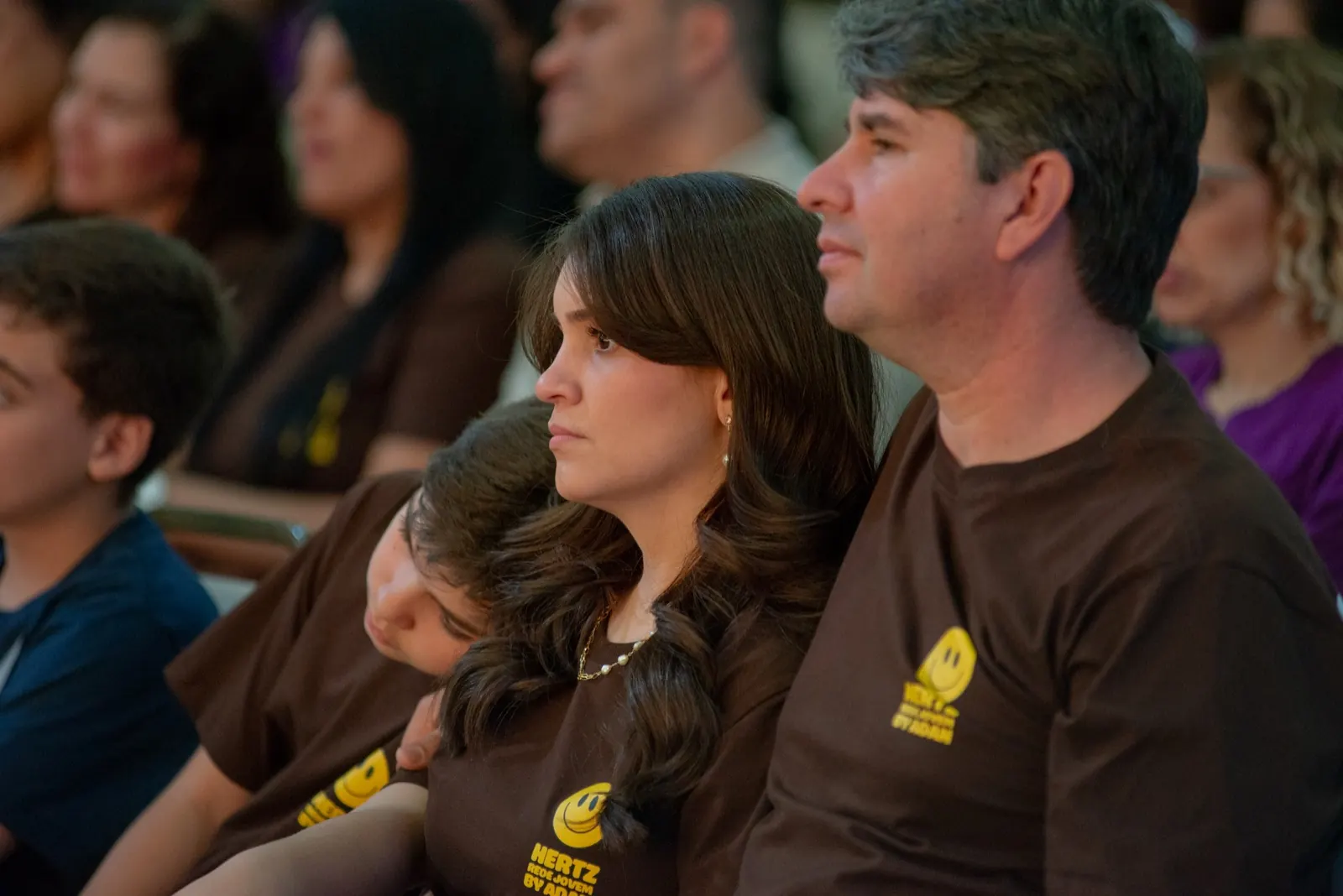 Família de camiseta marrom assistindo à programação, com um adolescente encostado em um dos pais